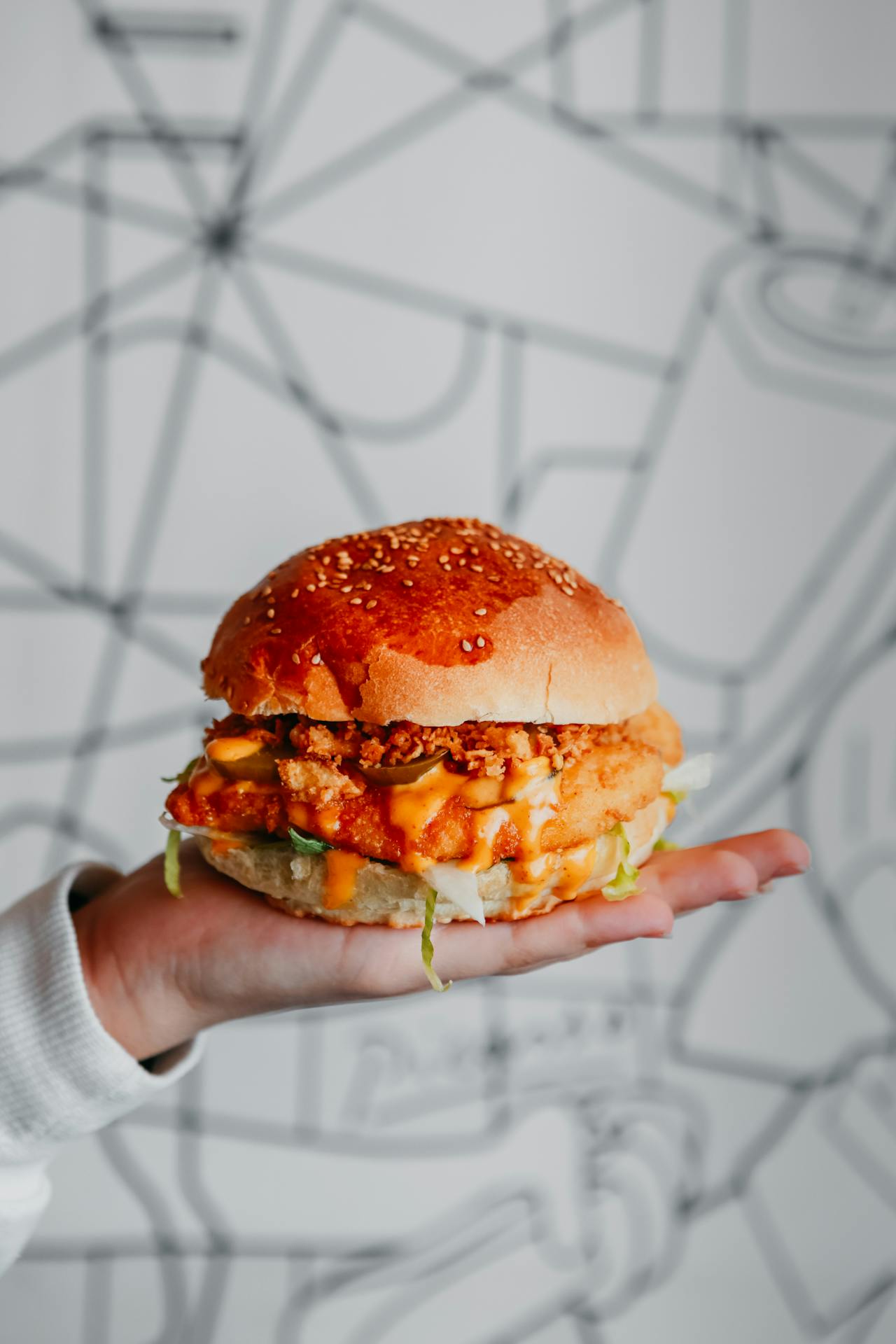 A hand holding up a crispy chicken burger drizzled with bright orange chipotle mayo, topped with pickled jalapeños and crispy panko crumbs on a sesame seed bun.