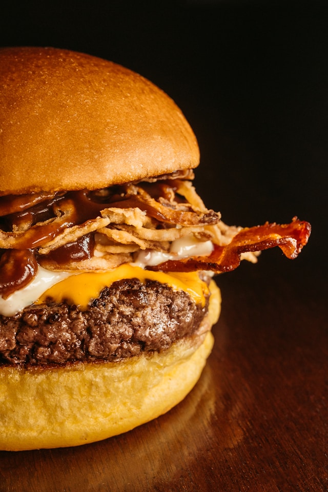A close-up of a thick cheeseburger with melted American cheese, crispy bacon, crispy fried onions, and A1 steak sauce dripping down the side on a soft brioche bun.