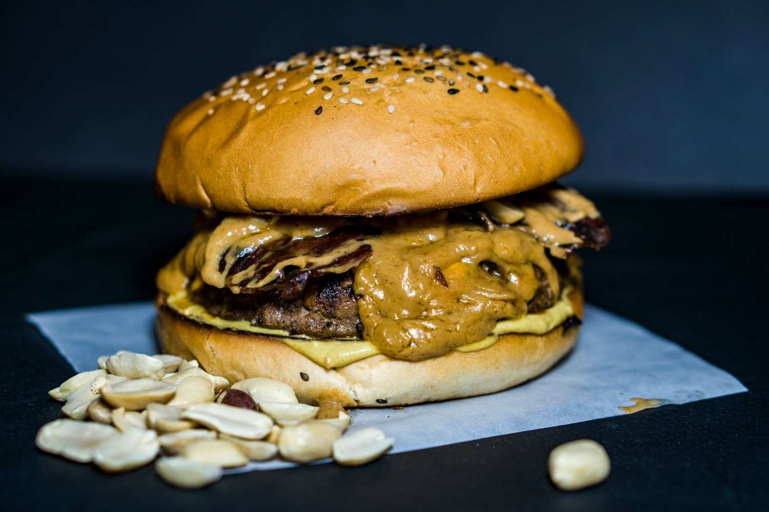 A PB&J-style peanut butter burger on a seeded sesame bun with a thick layer of creamy peanut butter, crispy bacon, and melted cheese, surrounded by scattered peanuts on a dark surface.