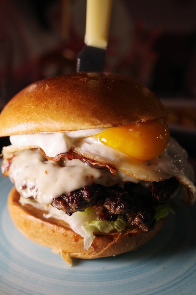 A breakfast burger topped with a sunny-side-up fried egg with a bright runny yolk, crispy bacon, and melted cheese on a toasted brioche bun with a knife stuck through the top.