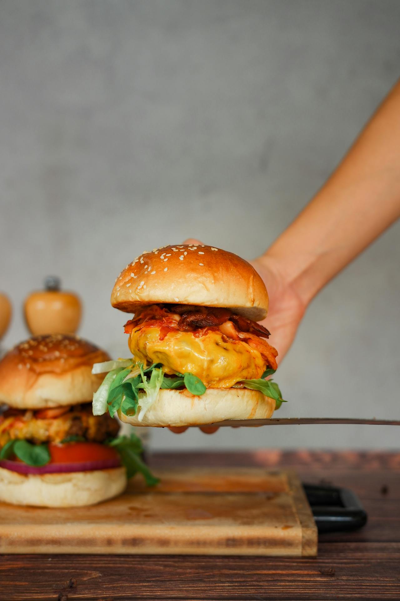 A hand holding up a fusion cheeseburger topped with spicy red kimchi, melted American cheese, bacon, and microgreens on a toasted sesame bun, with a second burger visible on a wooden board.