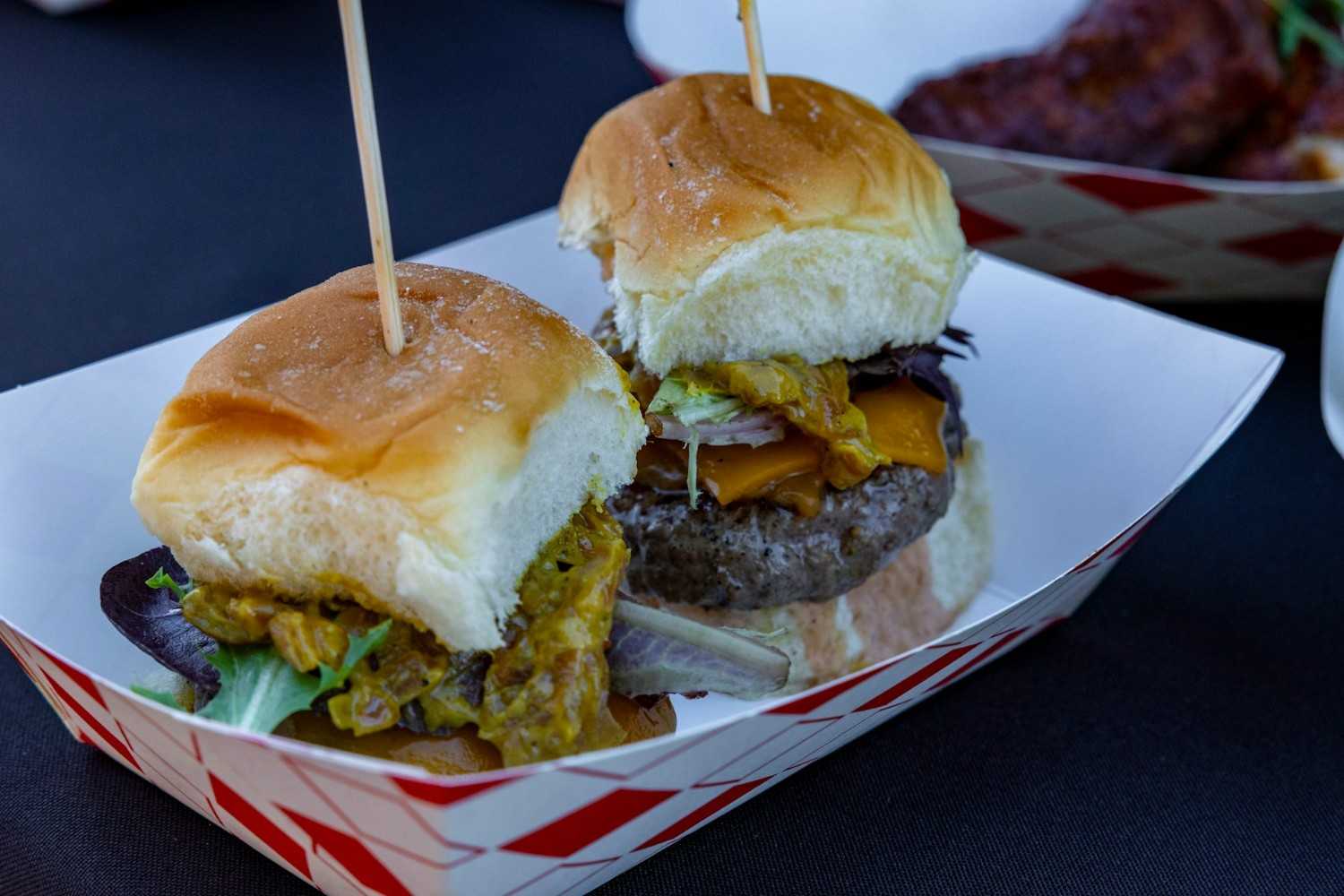 Two mini New Mexico-style green chile cheeseburgers on soft slider buns topped with roasted green chiles, melted cheddar, and mixed greens, served in a red and white check paper tray.