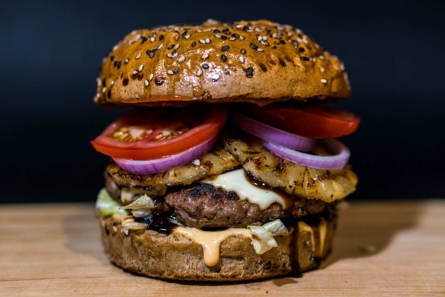 A tall Hawaiian-style burger on a seeded bun stacked with a charred grilled pineapple ring, teriyaki glaze, red onion rings, tomato, lettuce, and melted Swiss cheese on a wooden cutting board.