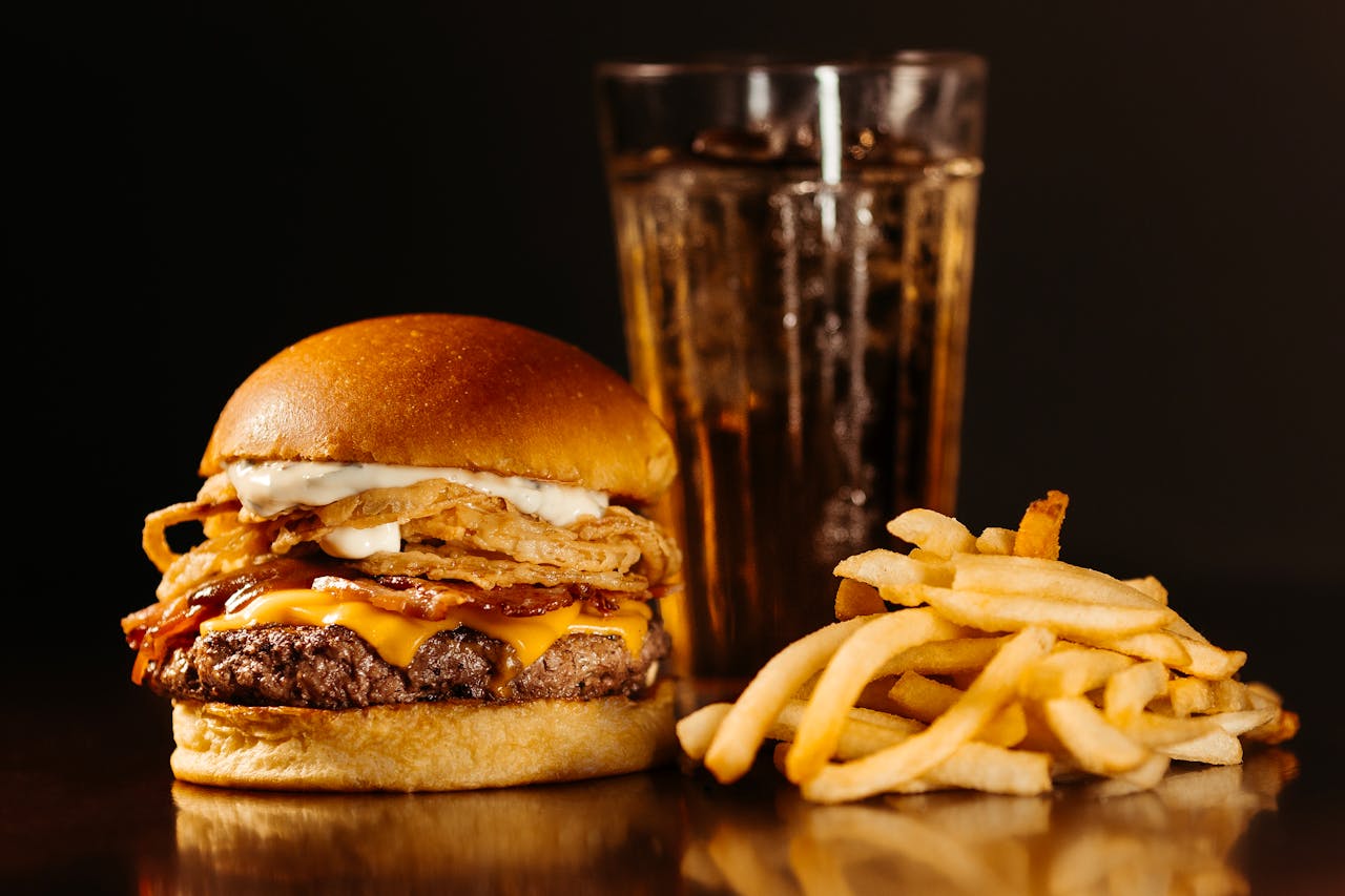 A classic American bacon cheeseburger topped with a generous pile of crispy fried onion straws and a dollop of aioli on a toasted brioche bun, served with fries and an iced drink.