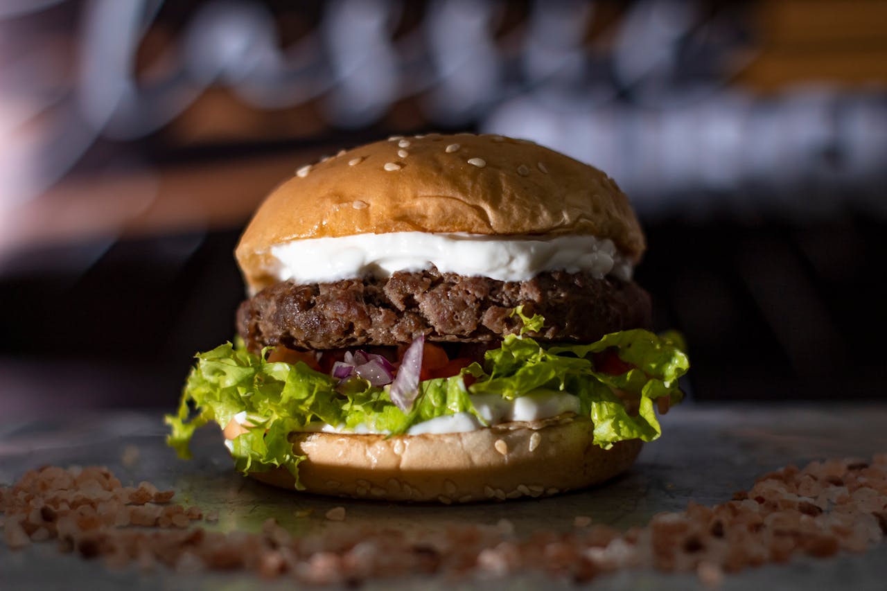 A Greek-style burger topped with thick, creamy tzatziki sauce and flecks of red onion, tomato, and curly lettuce on a sesame seed bun, with a grain scattered around the base.