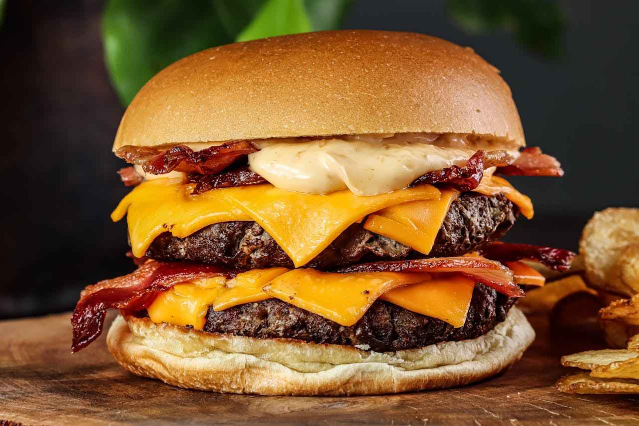 A towering double bacon cheeseburger with two thick beef patties, four slices of melted cheddar cheese, crispy bacon, and a drizzle of aioli on a soft brioche bun on a wooden board.