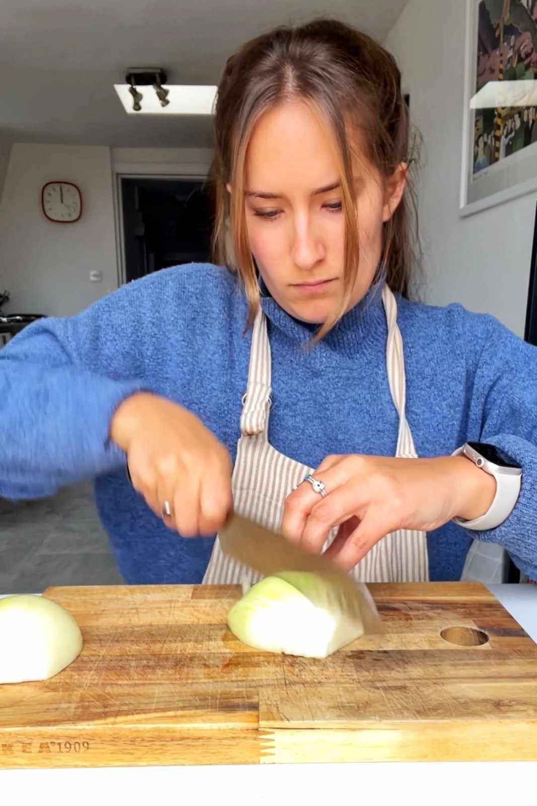 Emily slicing a white onion finely on a wooden cutting board with a chef's knife, wearing a striped apron.