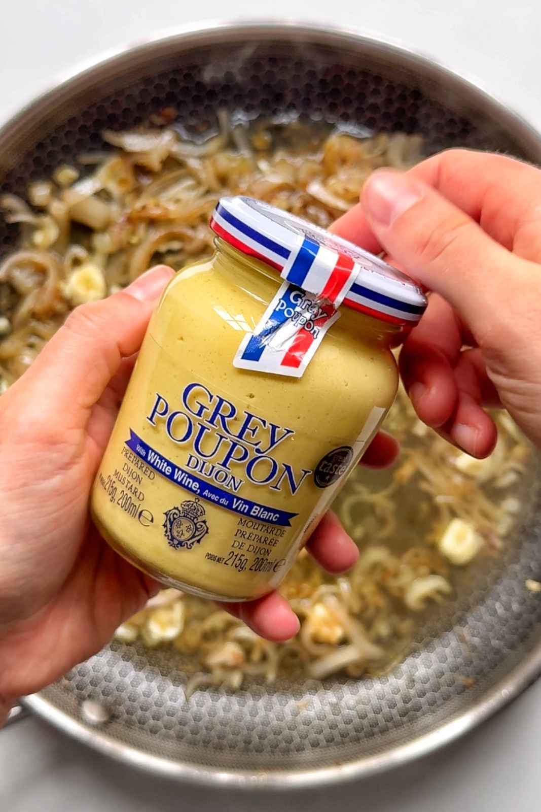 A jar of Grey Poupon dijon mustard about to be spread on top of a burger.