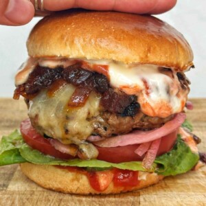 Close-up of a homemade cheeseburger on a toasted brioche bun with melted cheese, crispy bacon, caramelized onions, pickled red onion, tomato, lettuce, and burger sauce dripping down the sides.