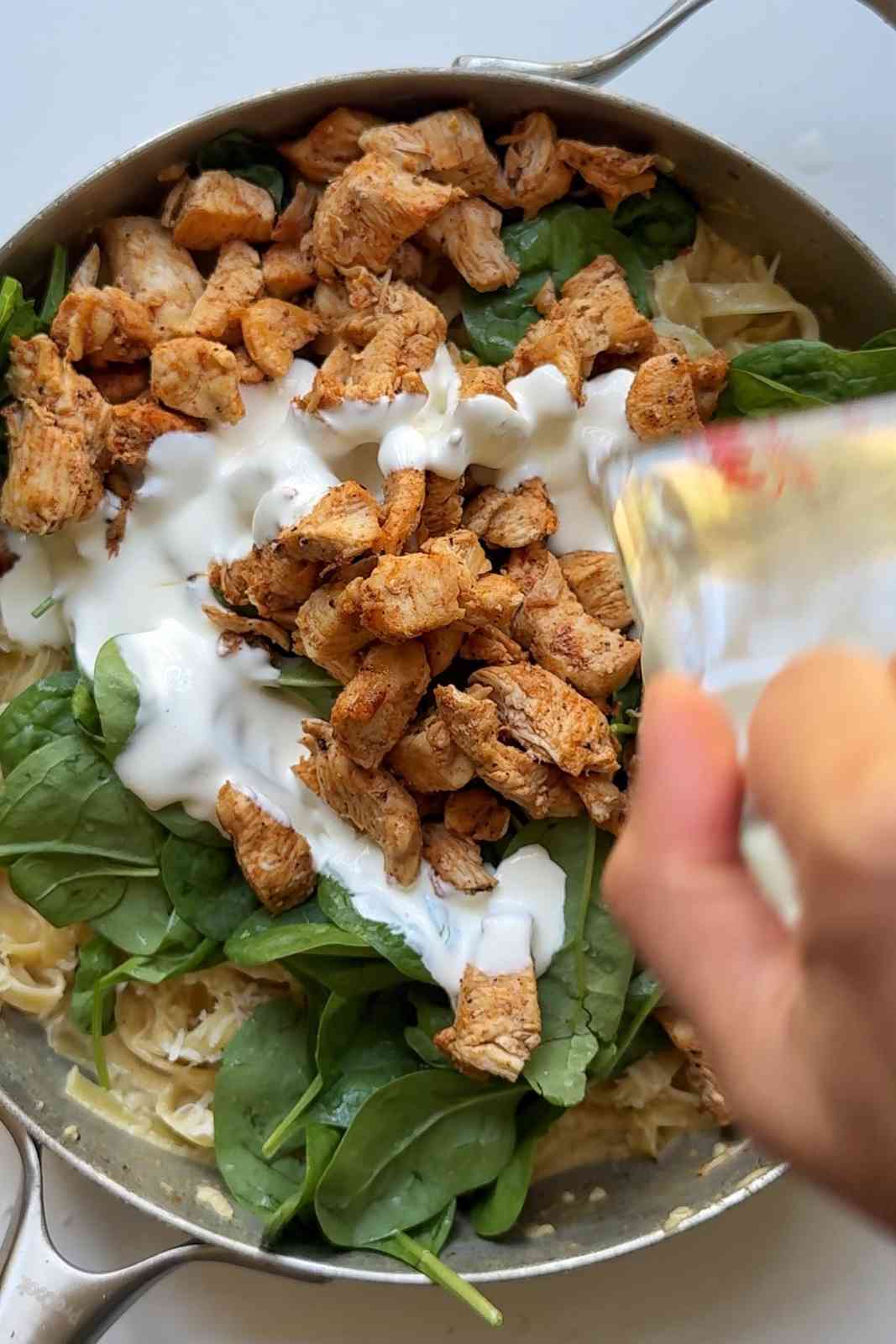 Pouring cream over chicken spinach alfredo pasta.