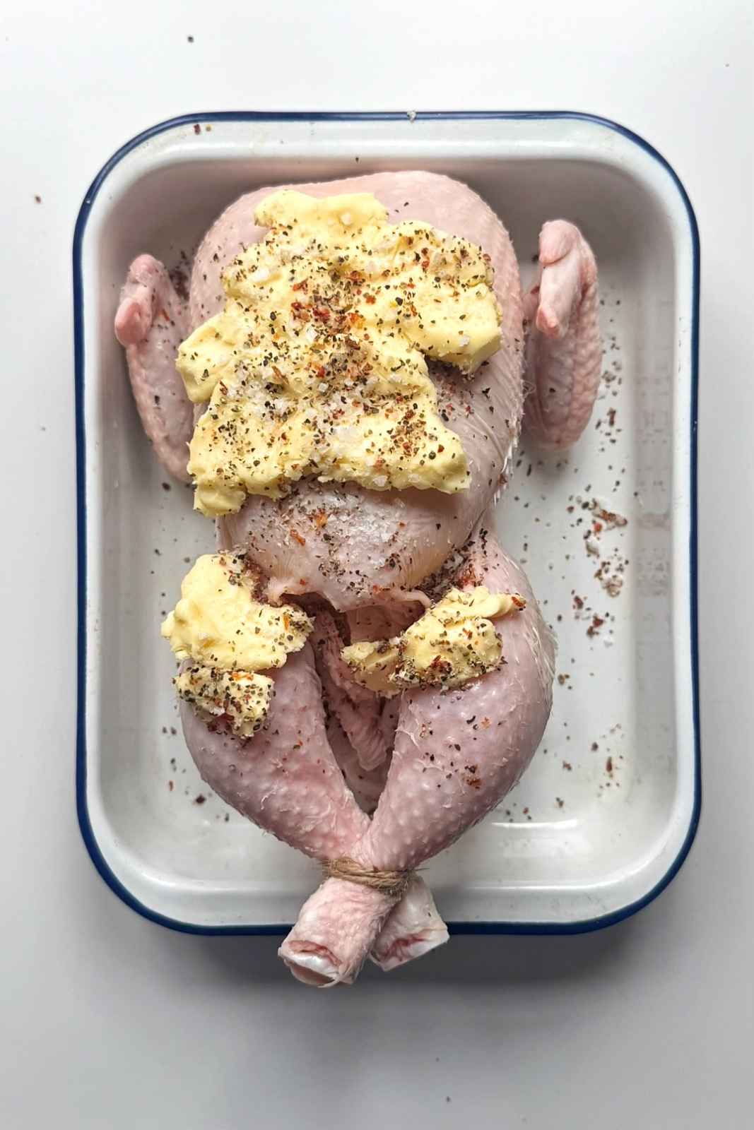 Raw whole chicken covered in butter and seasonings in a baking tray.