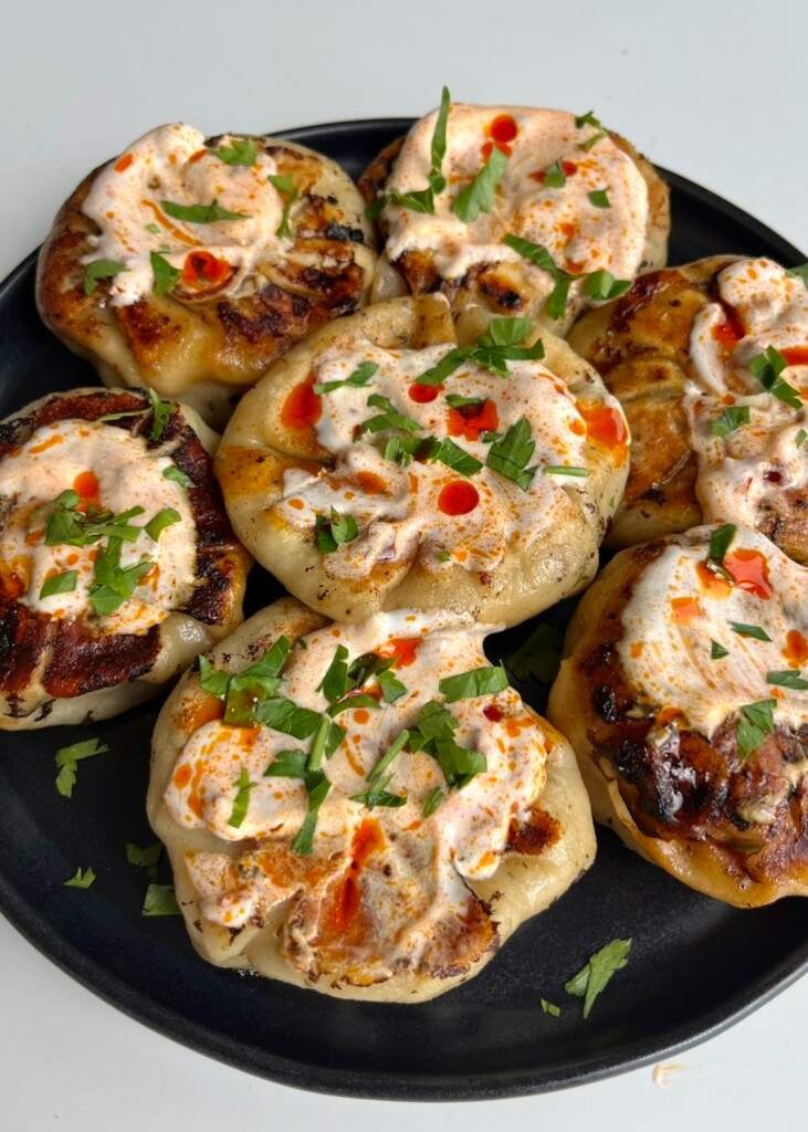 A black plate of pan-fried turkey dumplings, each topped with a creamy white sauce, drops of red chili oil, and a generous sprinkle of freshly chopped flat-leaf parsley.