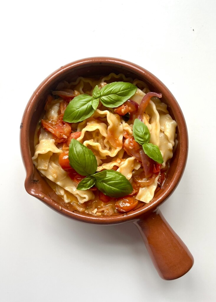 An overhead shot of wavy broad pasta in a roasted cherry tomato and red onion sauce, served in a rustic brown terracotta bowl with a handle against a white background, garnished with four vibrant, fresh basil sprigs.
