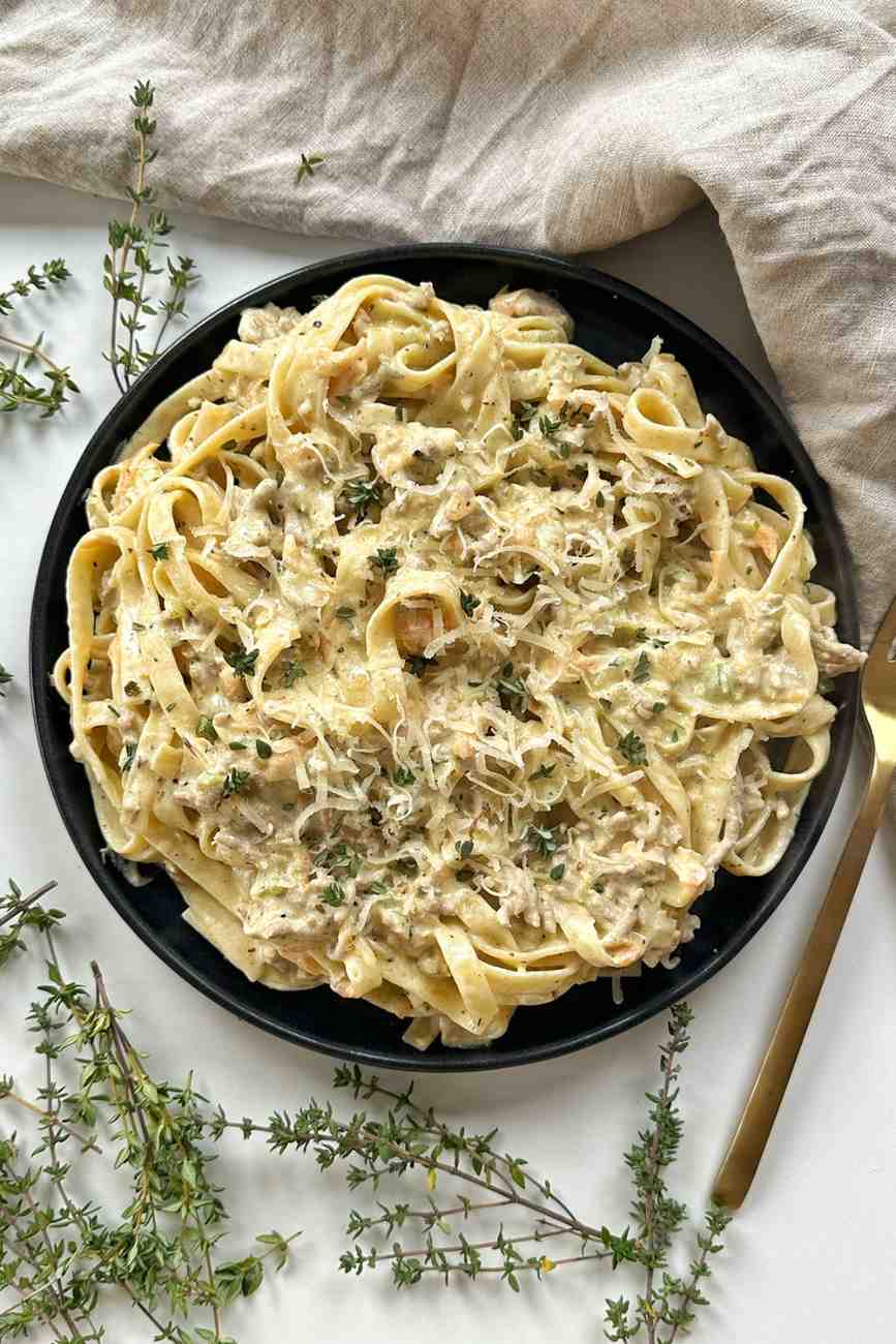 A top-down view of a black plate piled with creamy white ragu pasta, topped with grated cheese and fresh thyme leaves. Several sprigs of fresh thyme are scattered on the white surface around the plate.