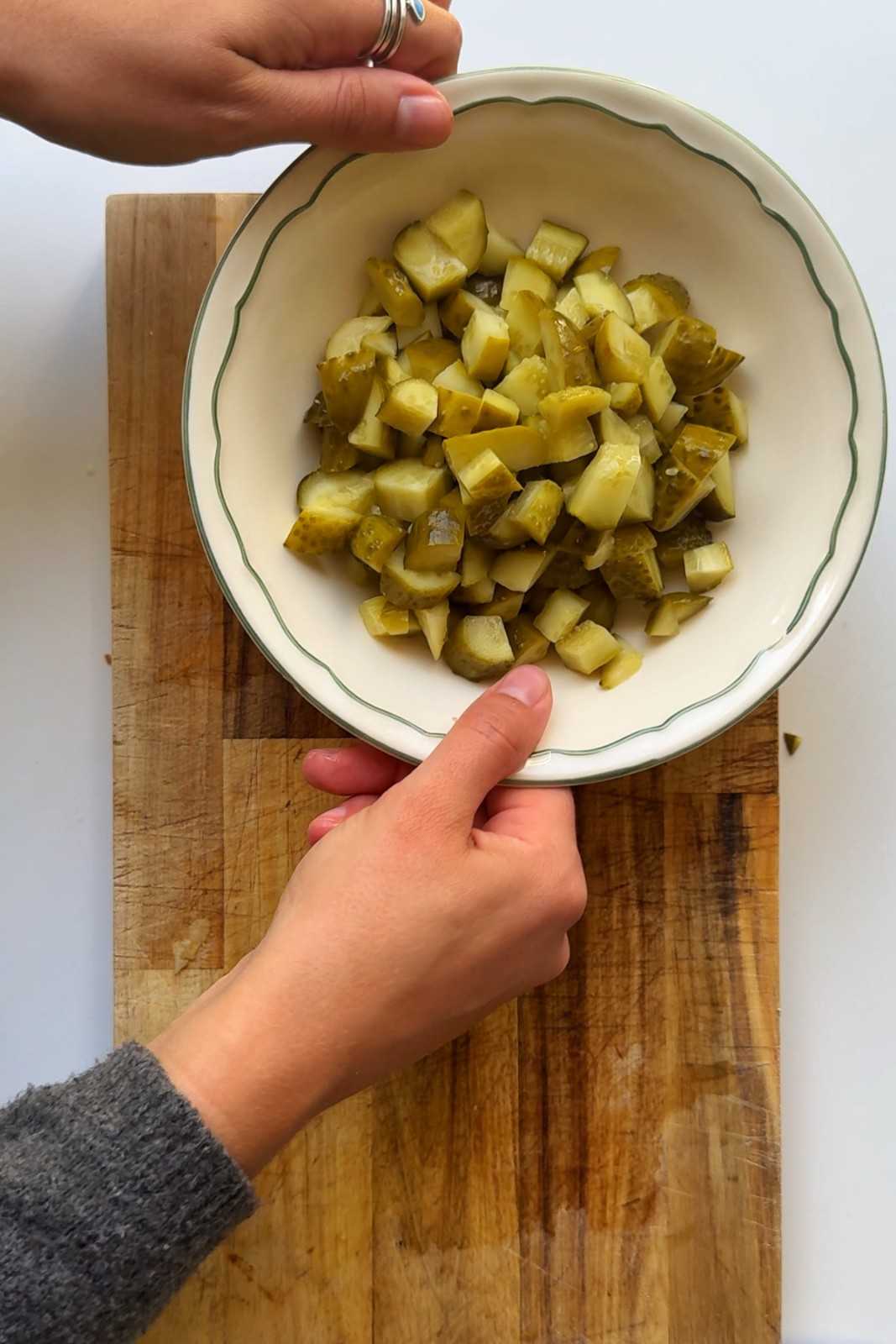 A top-down shot of a pair of hands holding a cream-colored bowl filled with diced green pickles over a rustic wooden cutting board.