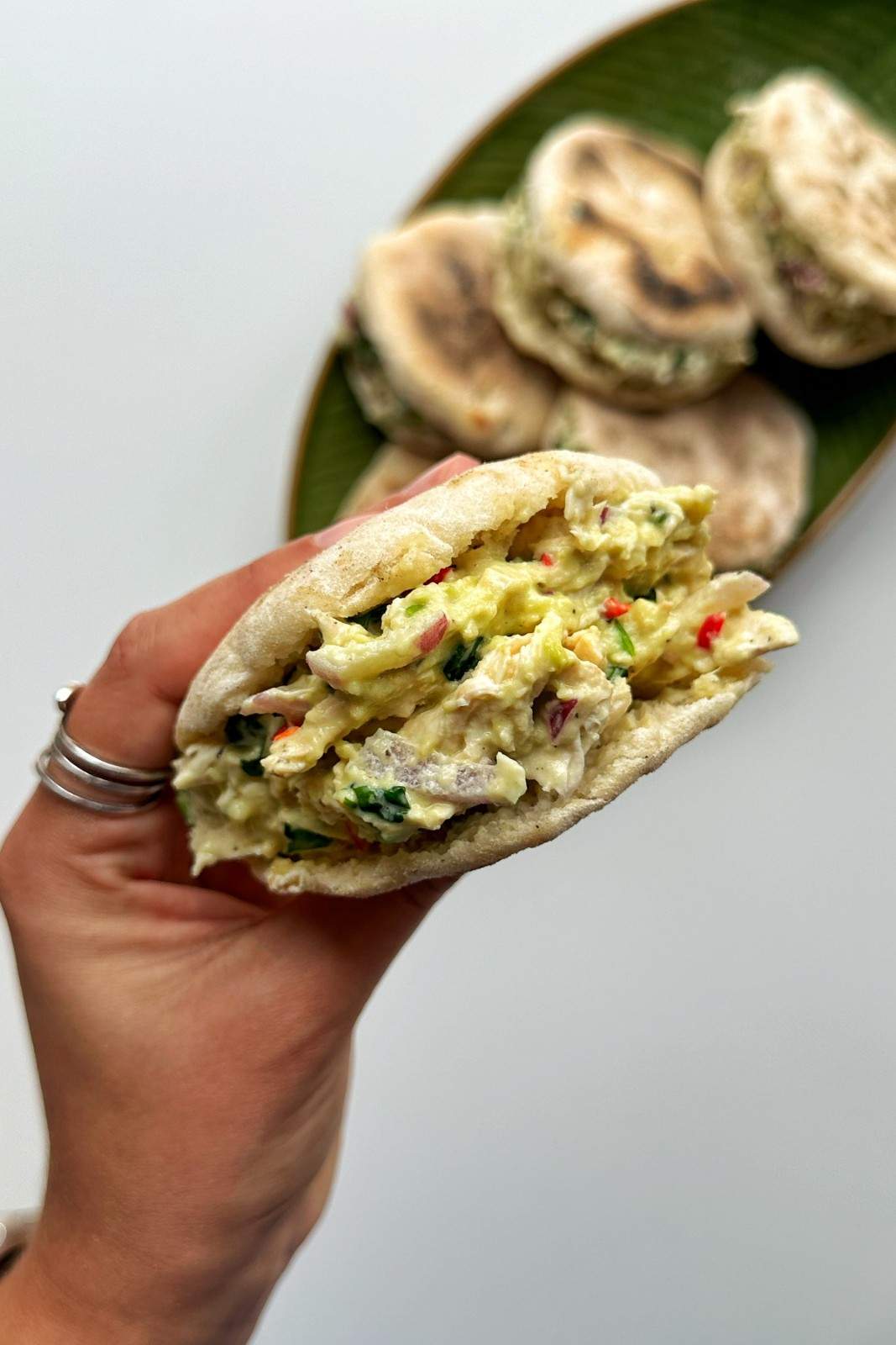 Cheat arepa held in hand showing creamy chicken and avocado filling with more arepas on a green leaf plate behind.