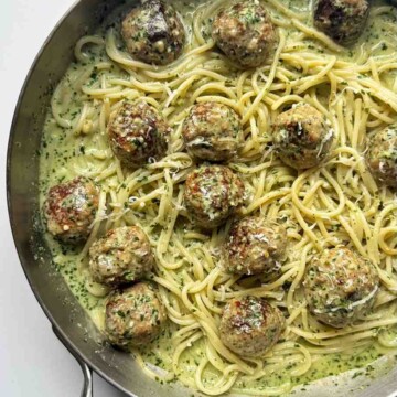 Pesto pasta and meatballs in a stainless steel skillet on a white background.