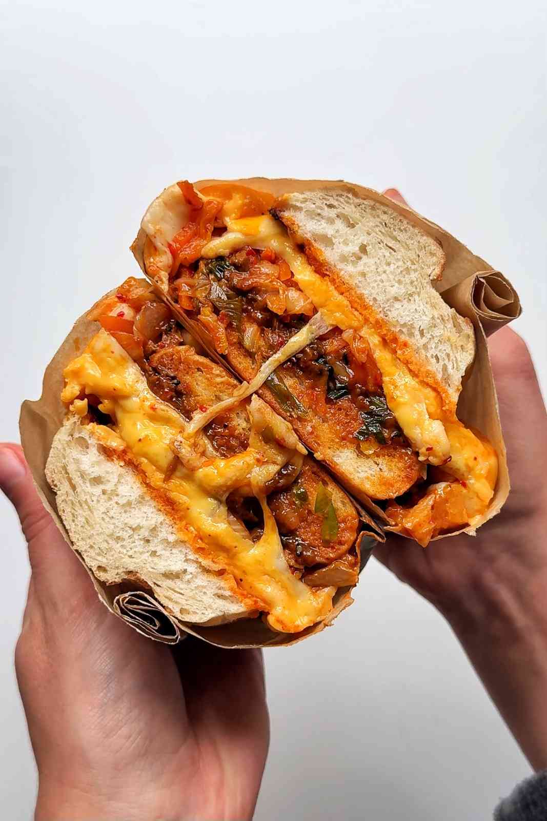Two hands holding a bulgogi cheesesteak sandwich wrapped in brown parchment paper, split open to reveal melted American and mozzarella cheese, stir-fried Korean bulgogi beef, kimchi, and spring onions in a white bread roll.