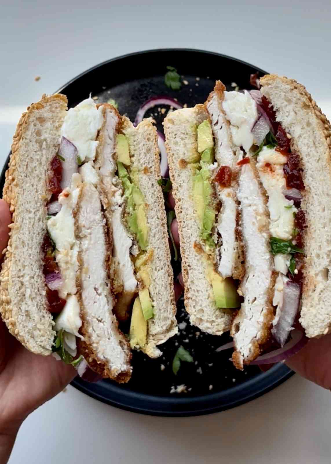 Two hands holding a Mexican cemita sandwich split in half over a black plate, showing layers of breaded chicken cutlet, sliced avocado, Oaxaca cheese, chipotle chilli, red onion, and cilantro in a seeded sesame bread roll. One of the least drippy sandwich recipes.