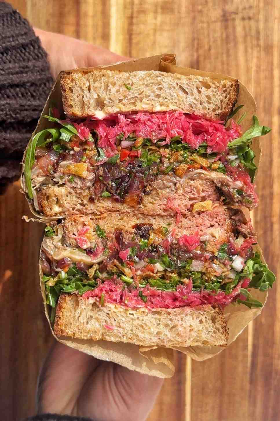 A hand holding a slow roast lamb sandwich wrapped in brown parchment paper, sliced in half to reveal layers of pulled lamb, fresh chimichurri, pickled beetroot, arugula, and deeply caramelised onions on toasted sourdough bread.