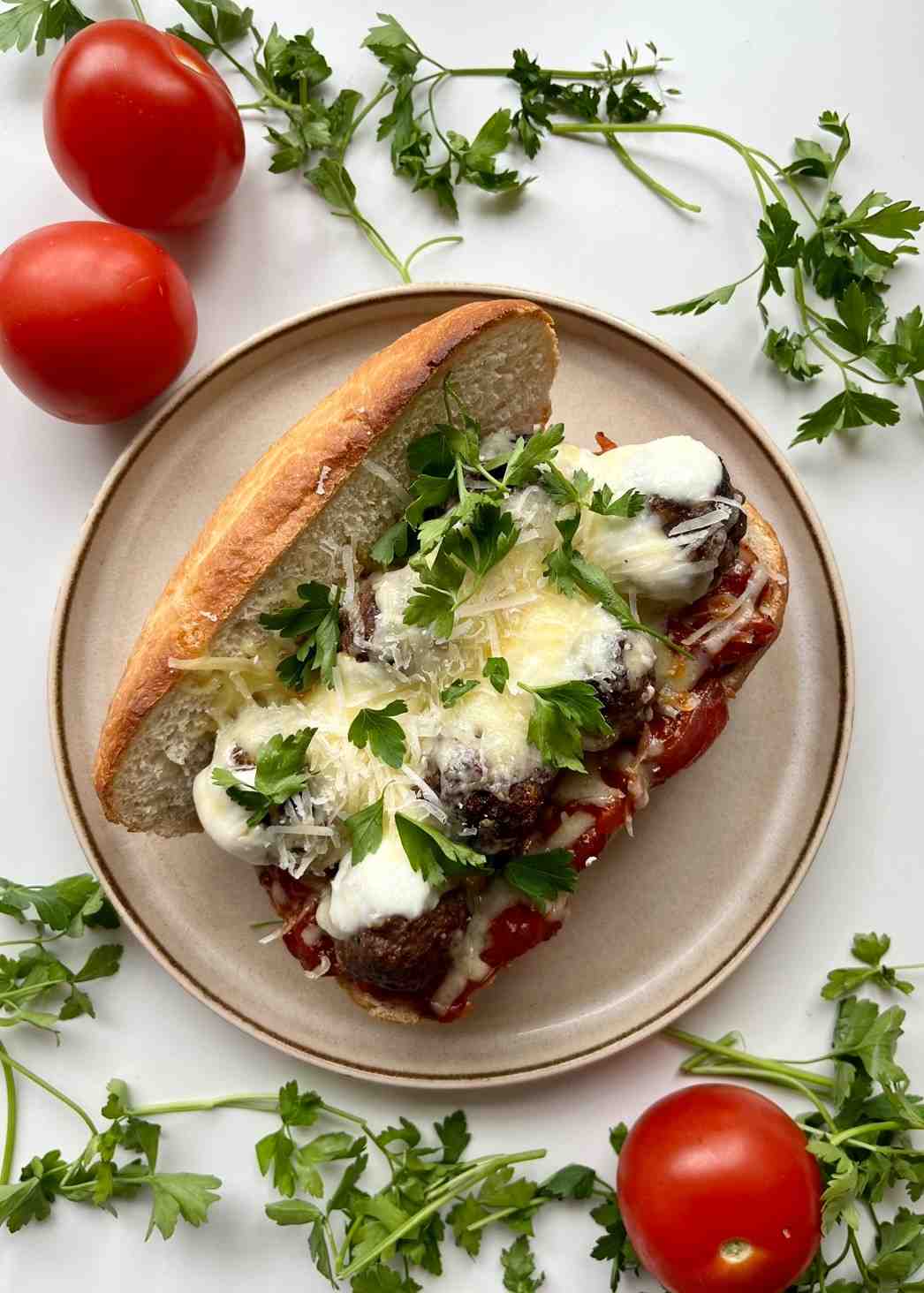 An open meatball sandwich on a white plate with homemade beef meatballs in marinara sauce, melted mozzarella, grated cheddar, and fresh parsley in a toasted sub roll, surrounded by fresh tomatoes and parsley on a white surface.