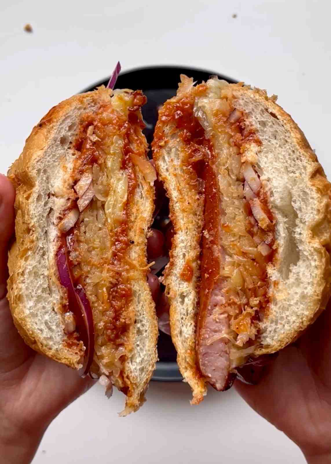 Two hands holding a Polish kielbasa sandwich split in half over a black plate, showing a butterflied smoked kielbasa sausage, sauerkraut, red onion, and homemade BBQ sauce in a crispy white bread roll.
