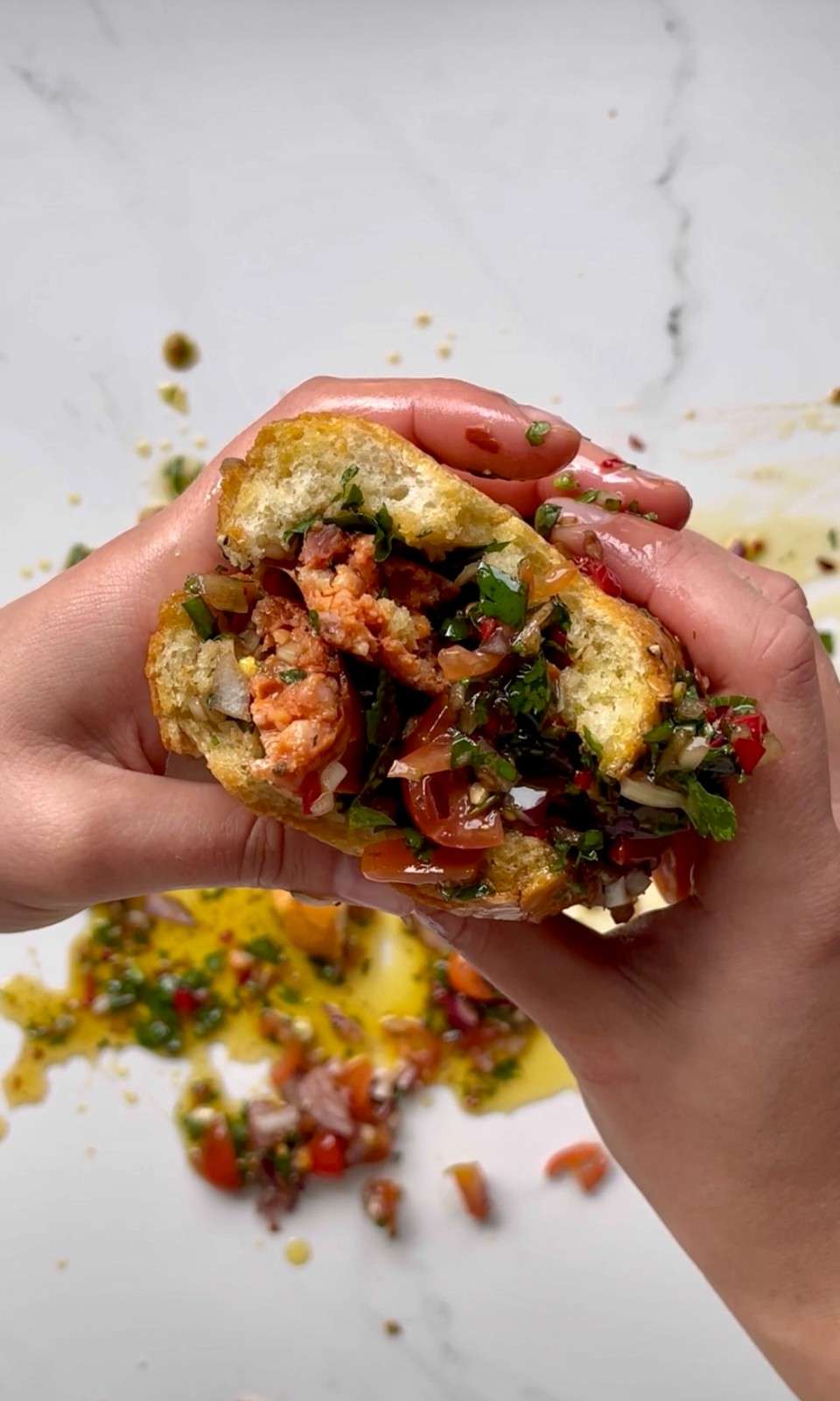 Two hands squeezing an Argentinian choripán sandwich, with grilled chorizo sausage, fresh chimichurri, and salsa criolla spilling out of a crusty white bread roll onto a marble surface.