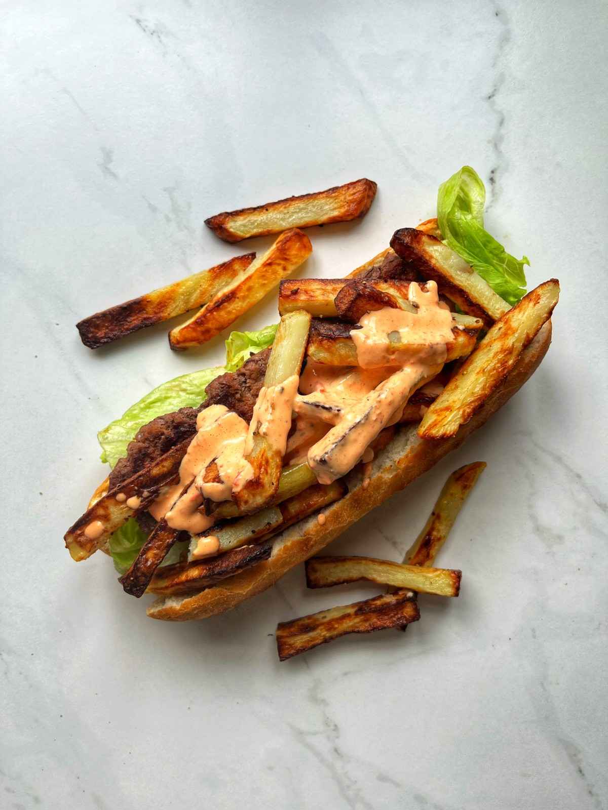 An open Belgian mitraillette sandwich on a marble surface, topped with a spiced beef patty, crispy homemade Belgian fries, lettuce, and a generous drizzle of homemade andalouse sauce spilling out of a baguette roll.