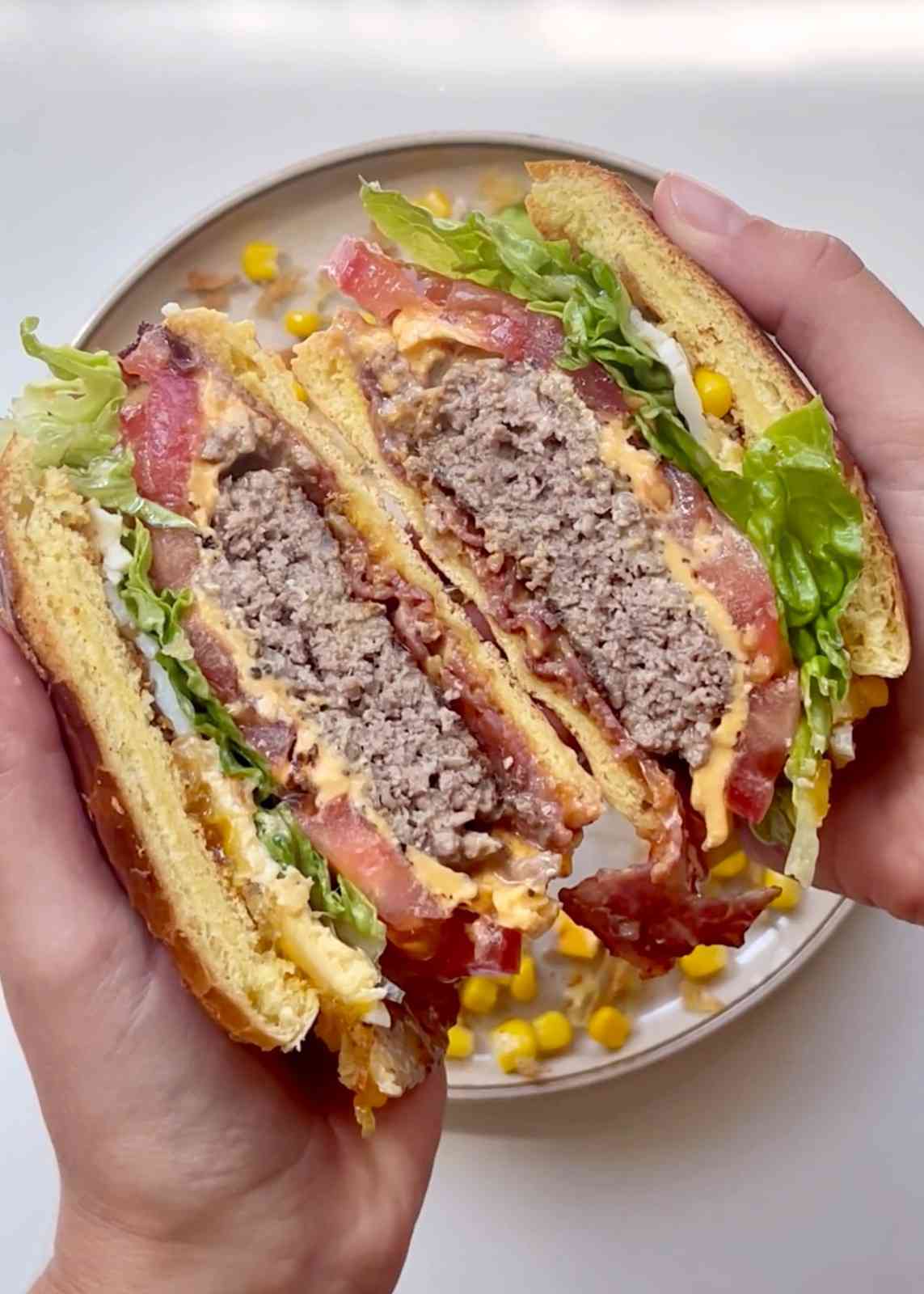 Two hands holding a Brazilian X-Tudo sandwich split in half over a plate, revealing layers of beef patty, bacon, fried egg, melted American cheese, sweetcorn, tomato, and lettuce with ketchup and mayonnaise in a toasted burger bun.