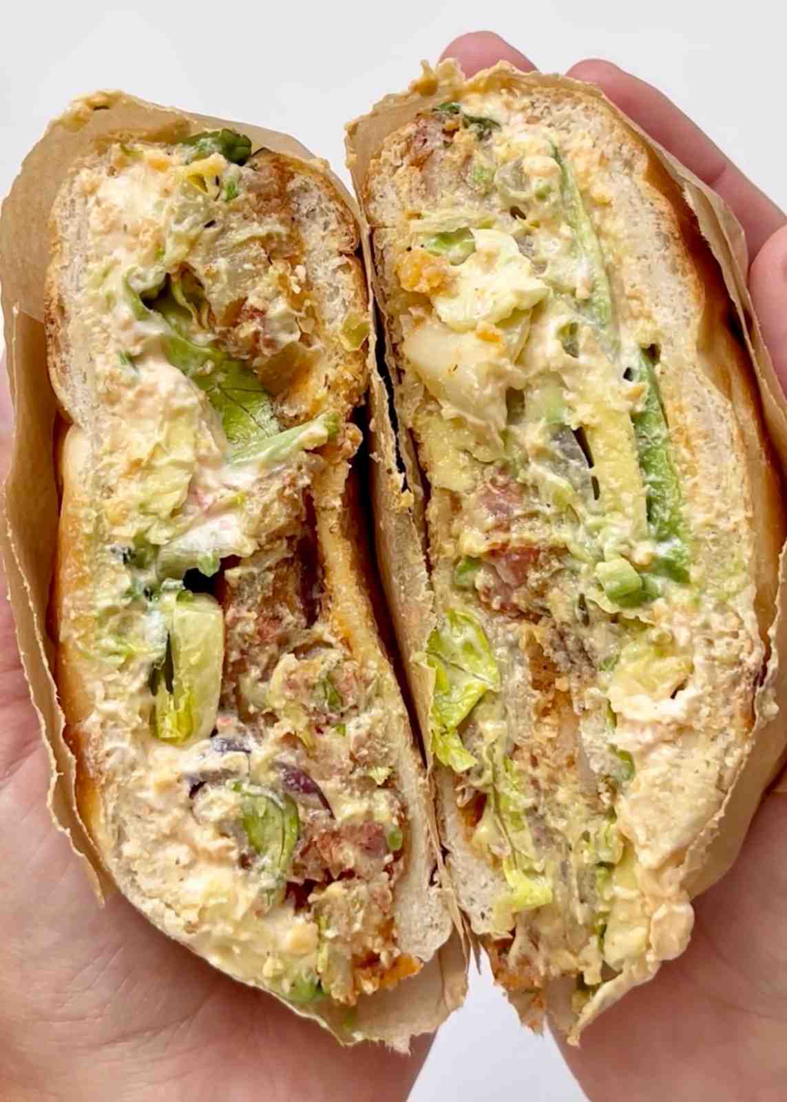 Two hands holding a Mexican pambazo sandwich split in half and wrapped in brown parchment paper, showing a filling of spiced chorizo and potato, creamy cheese, sour cream, avocado, and lettuce in a bread roll.