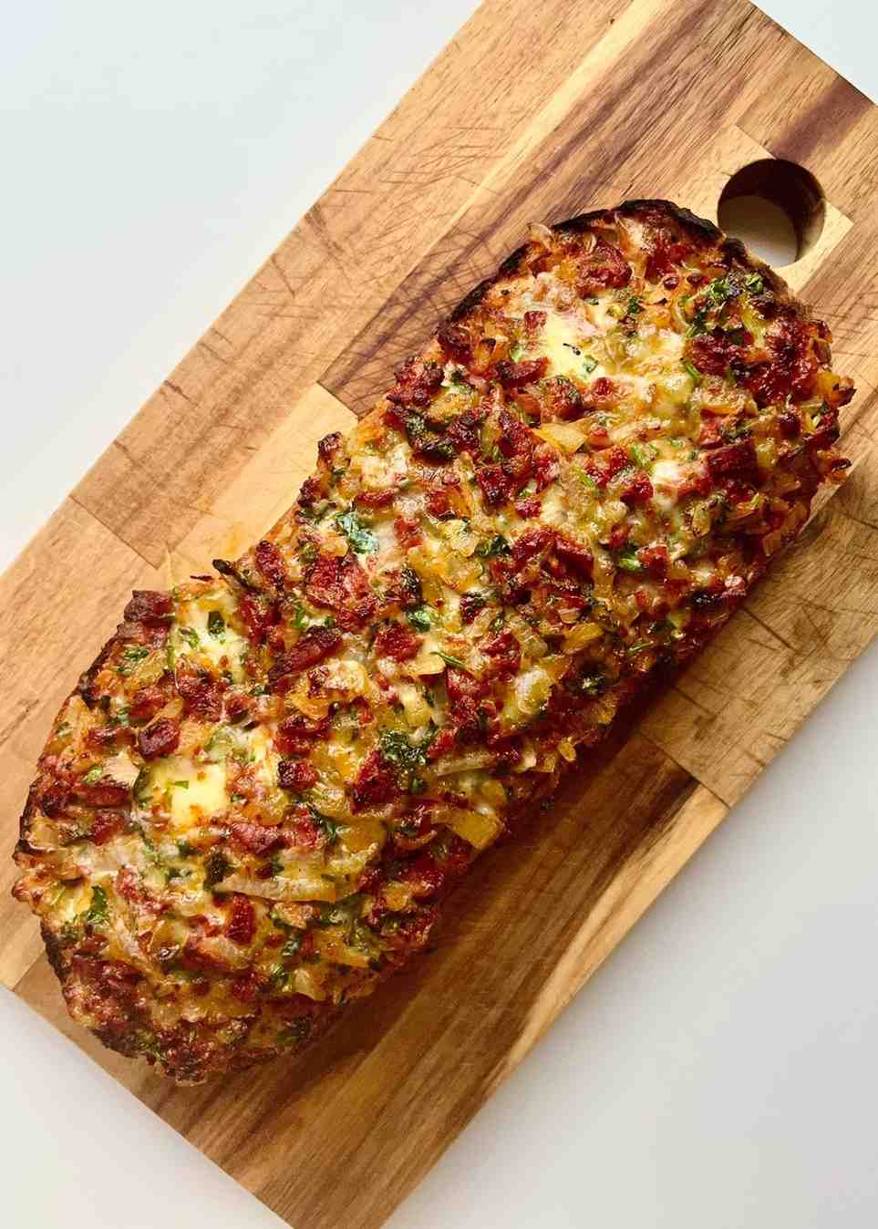 A cheesy 'nduja slider on a wooden chopping board, with a ciabatta loaf topped with a bubbling golden crust of melted mozzarella, chorizo, 'nduja paste, onion, garlic, and fresh parsley, grilled until crispy at the edges.