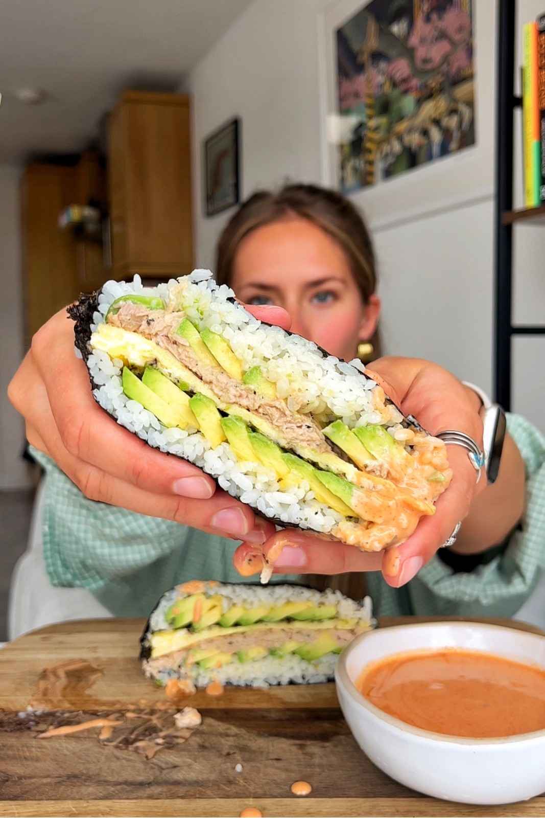 Emily holding up an onigirazu Japanese sushi sandwich sliced in half to show neat layers of tuna mayonnaise, omelette, and sliced avocado pressed between sushi rice and wrapped in nori, with a bowl of sriracha dipping sauce on a wooden board below.
