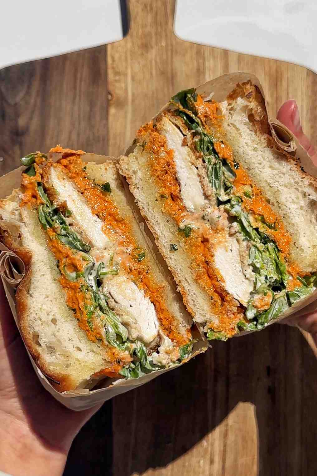 A hand holding a crunchy chicken romesco sandwich wrapped in brown parchment paper, sliced in half to show layers of panko-fried chicken cutlet, vibrant orange romesco sauce, and dressed arugula salad on focaccia bread, resting on a wooden chopping board.