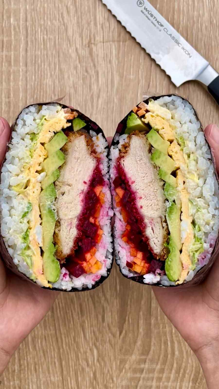 Two hands holding a katsu onigirazu sushi sandwich split in half to reveal vivid layers of breaded chicken katsu, sliced avocado, egg, pickled beetroot, grated carrot, and white cabbage pressed between sushi rice and wrapped in nori, with a knife on a wooden board above.