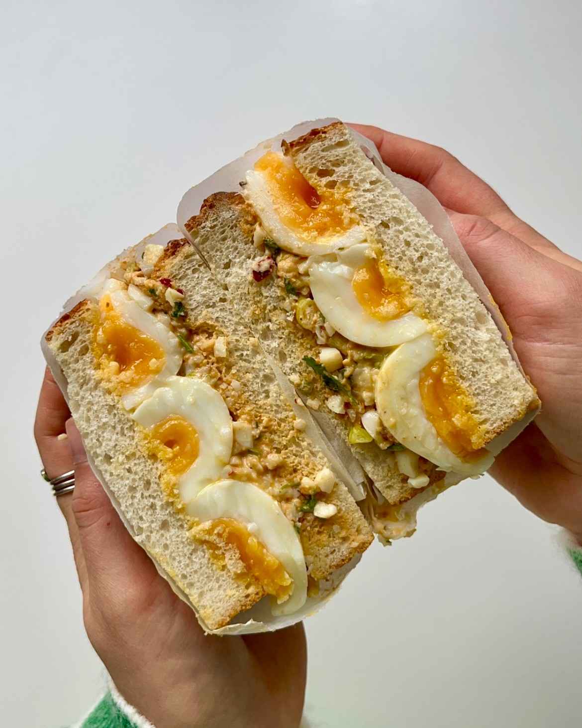 Two hands holding a tamago harissa sando sliced in half, showing jammy soft-boiled eggs with vivid orange runny yolks nestled in a creamy spiced harissa egg salad with spring onion and parsley between two slices of thick white bread.