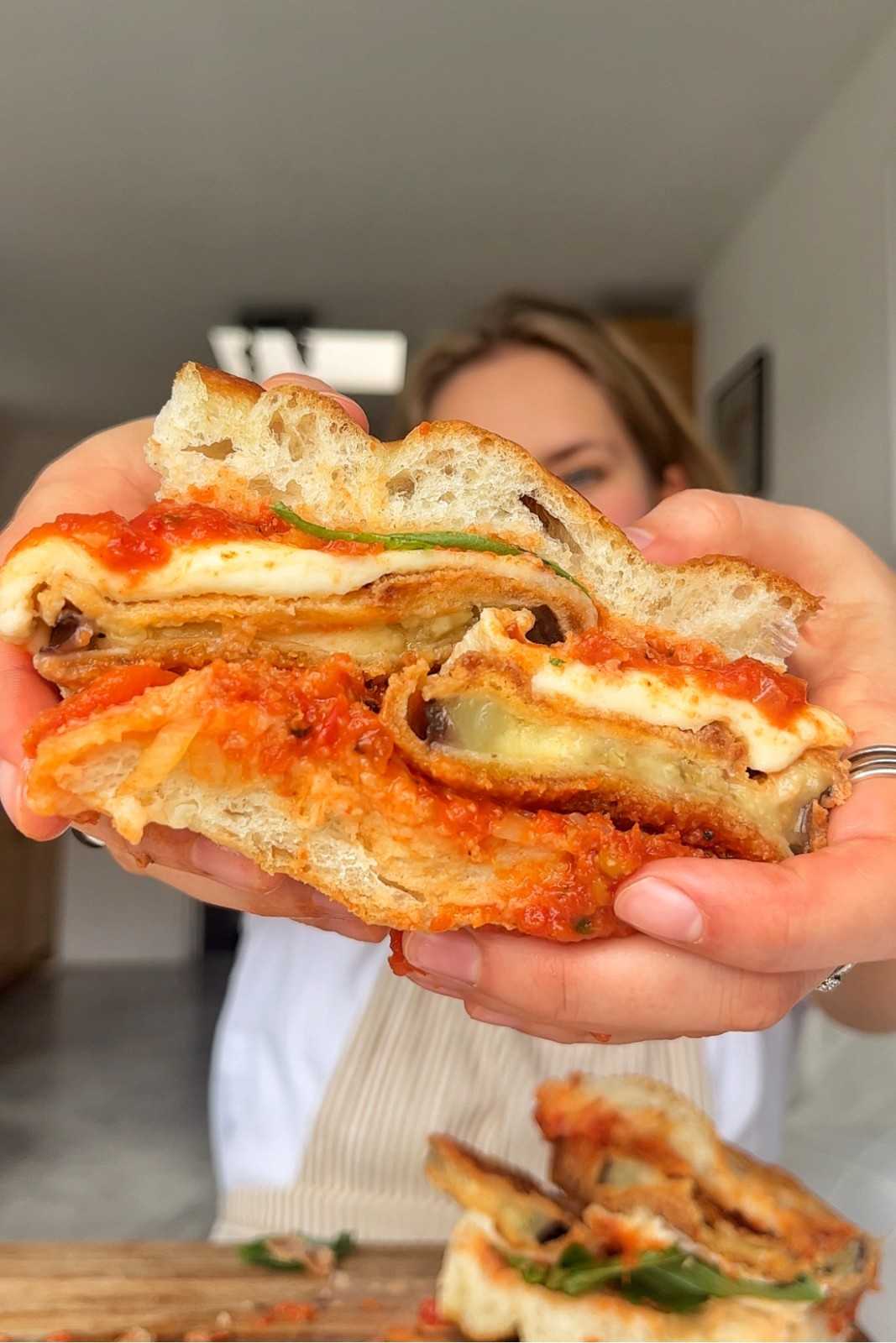 Emily holding up an eggplant parmesan sandwich sliced in half, showing layers of panko-fried eggplant discs, melted mozzarella, vibrant red marinara sauce, and fresh basil in toasted focaccia bread.