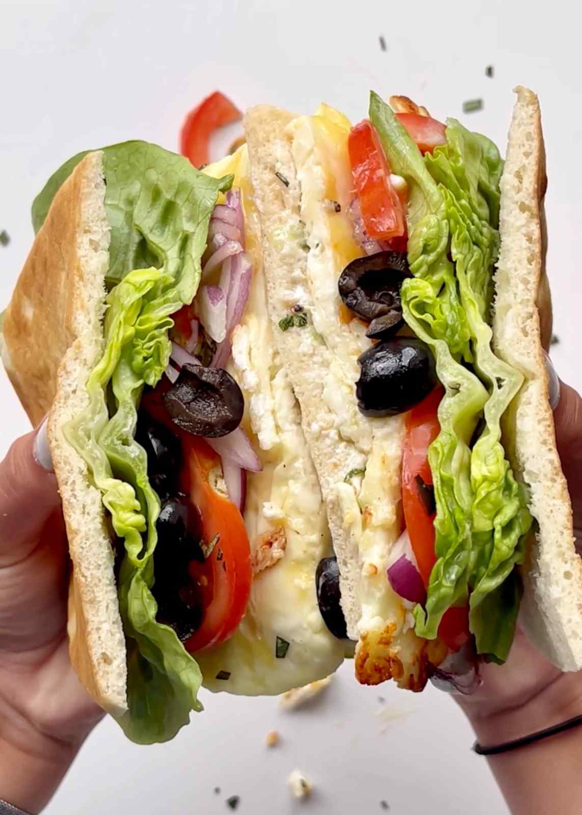 Two hands holding a Cypriot halloumi sandwich split in half, showing layers of pan-fried halloumi, fresh lettuce, sliced tomato, black olives, red onion, and creamy talatouri sauce in a thin focaccia bread roll.