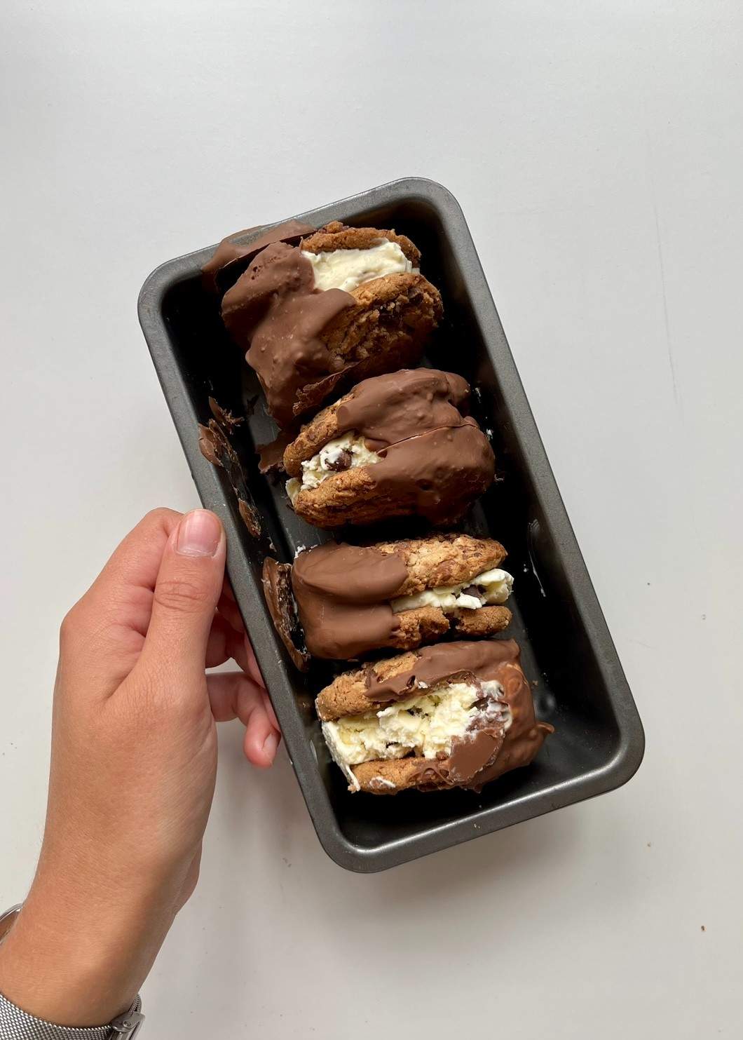 A hand holding a loaf tin containing four homemade ice cream sandwiches, each with a scoop of no-churn vanilla chocolate chip ice cream pressed between two chocolate chip cookies and half-dipped in melted chocolate.