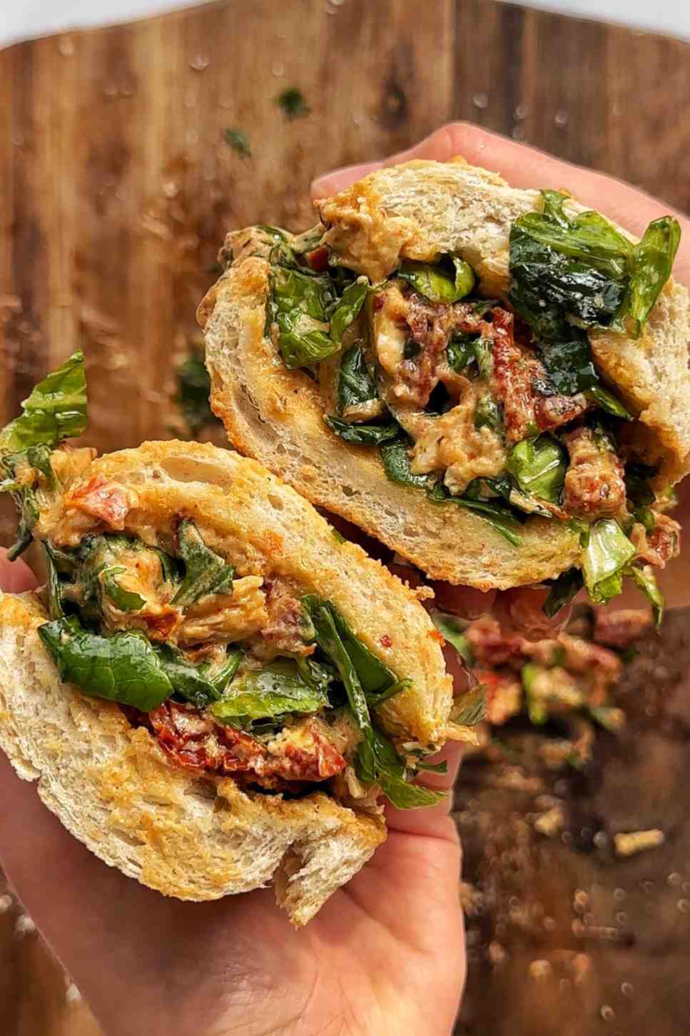 Two hands holding a marry me chicken chopped salad sandwich split in half, showing a generous filling of creamy sun-dried tomato chicken, fresh arugula, and basil chopped together and piled into a crusty baguette roll.