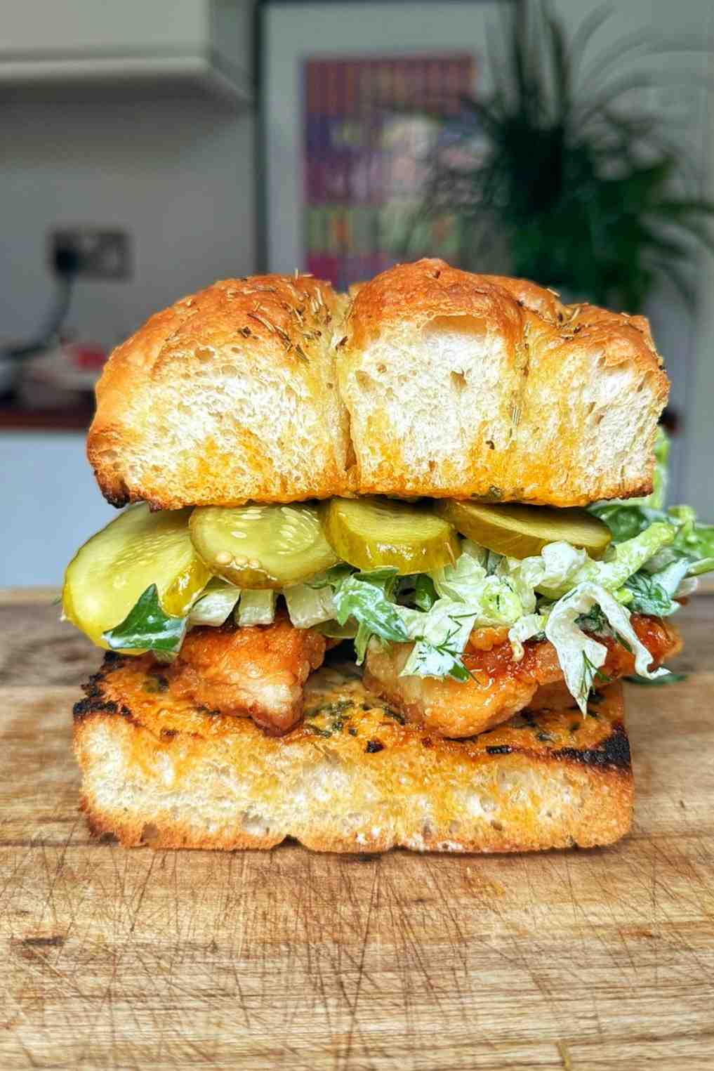 A hot honey chicken sandwich on toasted focaccia bread, layered with a golden fried chicken cutlet coated in hot honey oil, dill yogurt dressed lettuce, and sliced cucumber pickles, sitting on a wooden chopping board.