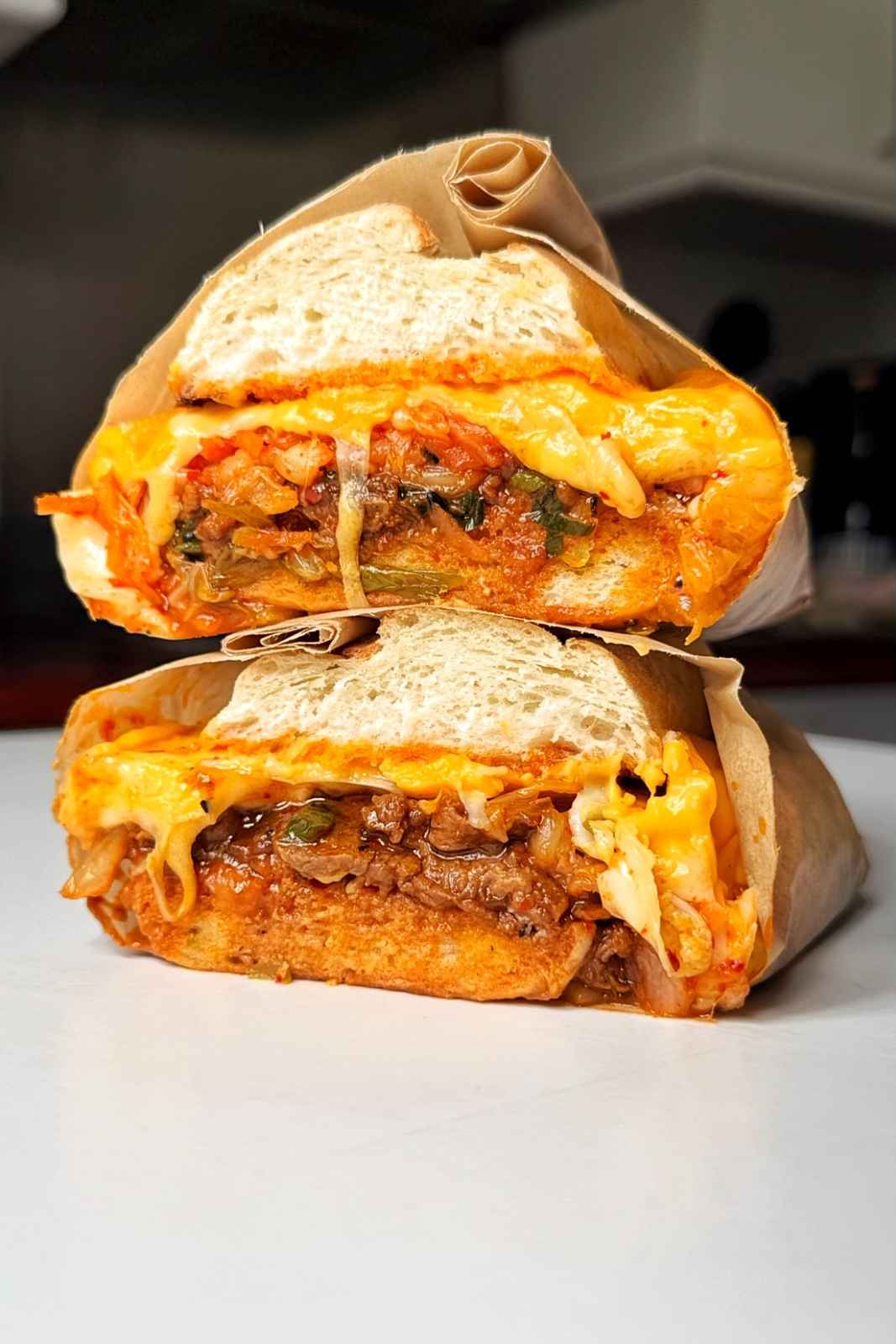 A bulgogi cheesesteak sandwich wrapped in brown parchment paper, sliced in half and stacked to show layers of thinly sliced marinated Korean beef, kimchi, spring onions, and melted American and mozzarella cheese oozing out of a white bread roll.