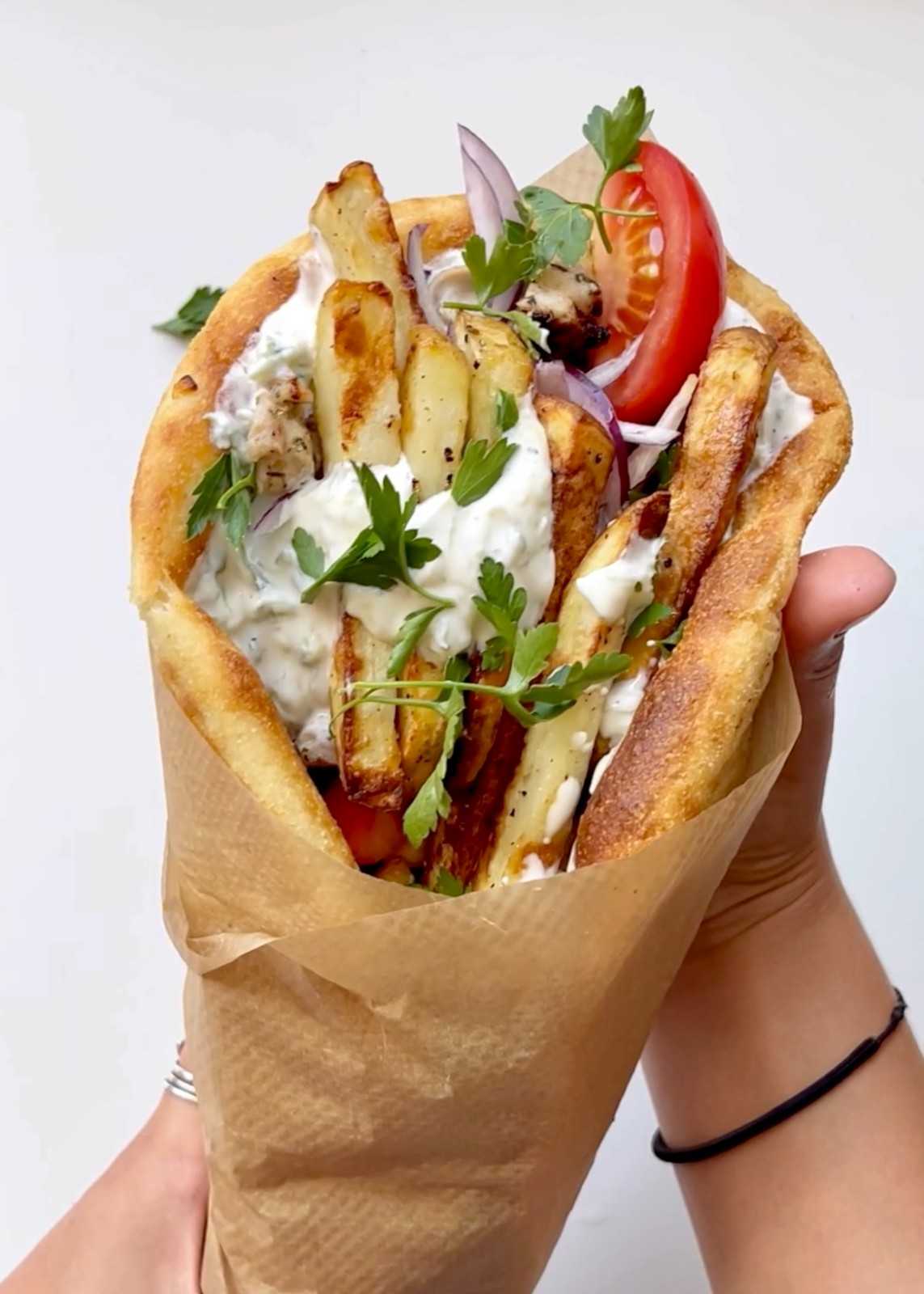 Two hands holding a Greek chicken gyros wrapped in brown parchment paper, filled with air fryer chicken, crispy homemade fries, generous tzatziki sauce, fresh tomato, red onion, and parsley in a toasted flatbread.