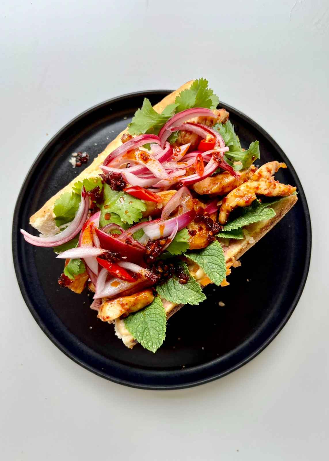 An open Vietnamese bánh mì baguette on a black plate topped with marinated chicken strips, pickled red onion, fresh mint leaves, cilantro, sliced red chilli, and a drizzle of chilli oil.