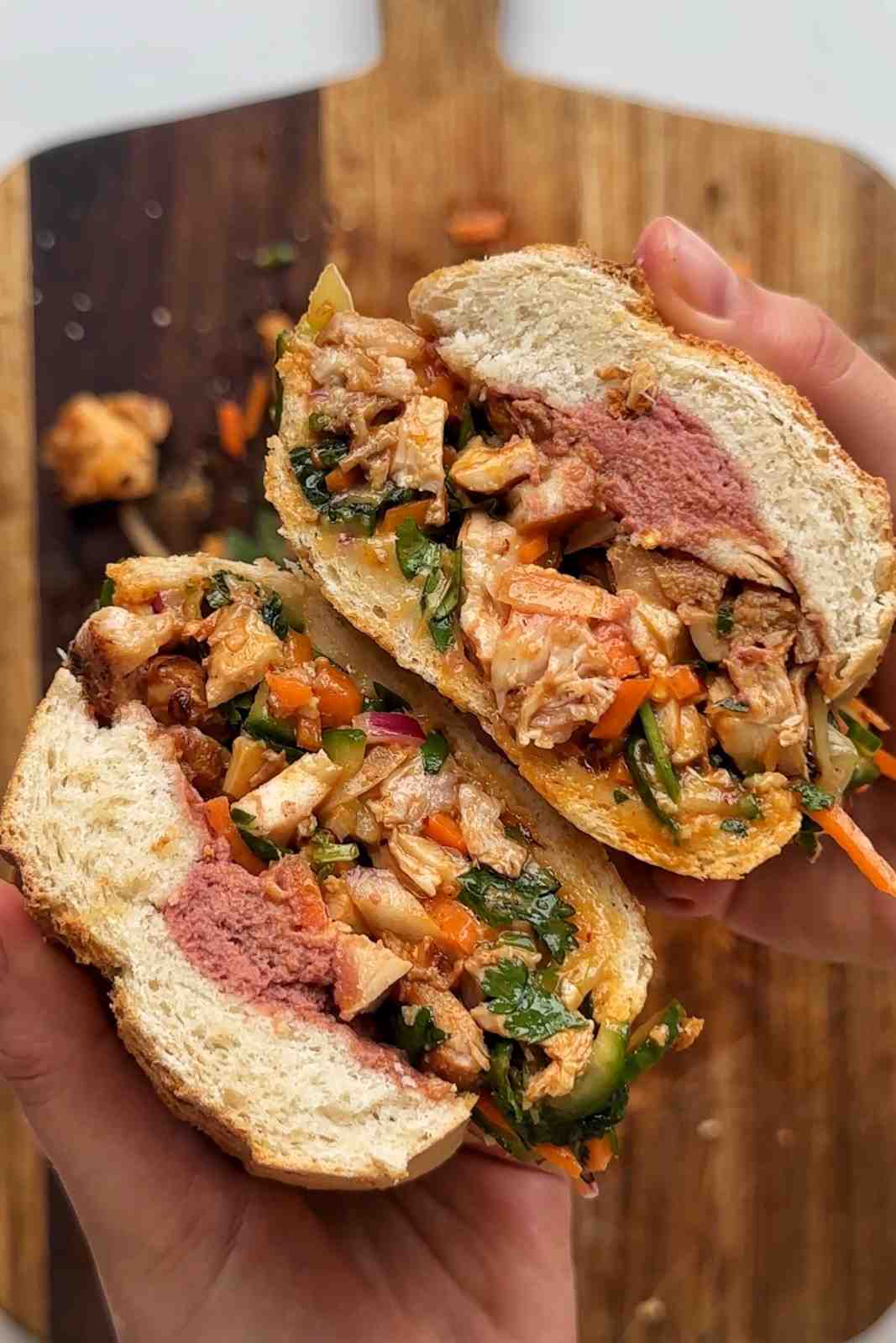 Two hands holding a bánh mì chopped salad sandwich split open over a wooden chopping board, packed with finely chopped fried marinated chicken, pickled carrot, cucumber, red onion, fresh cilantro, mint, and a swipe of pâté in a crusty baguette roll.