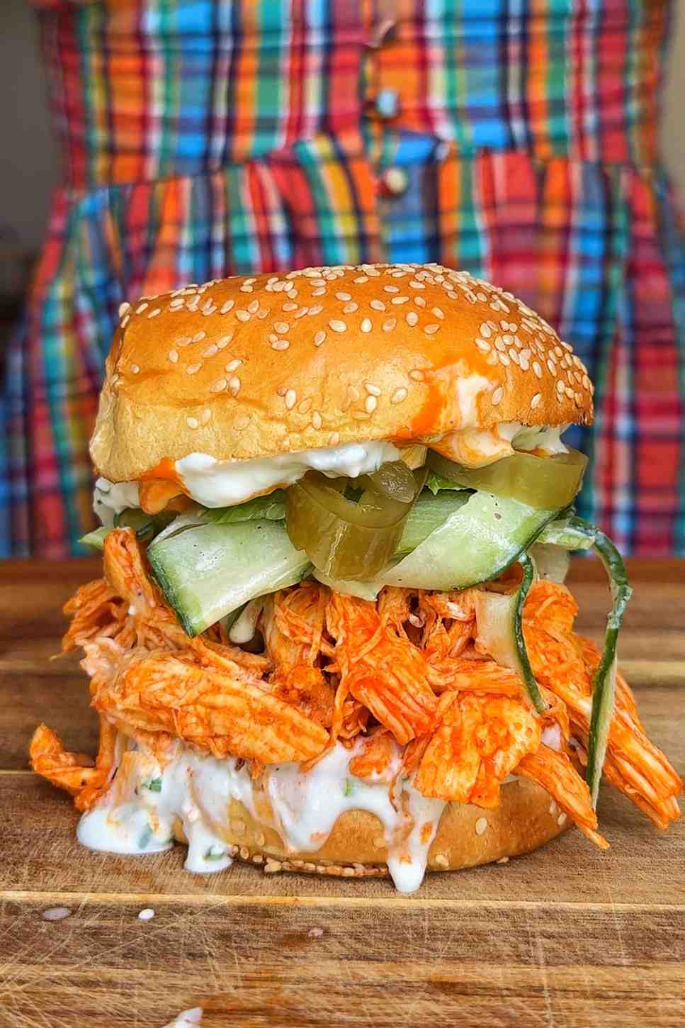 A shredded buffalo chicken sandwich on a toasted sesame brioche bun, piled high with bright orange buffalo-sauced shredded chicken, cucumber celery slaw, sliced jalapeños, and blue cheese sauce dripping down the sides, on a wooden chopping board.