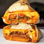 A bulgogi cheesesteak sandwich wrapped in brown parchment paper, sliced in half and stacked to show layers of thinly sliced marinated Korean beef, kimchi, spring onions, and melted American and mozzarella cheese oozing out of a white bread roll.