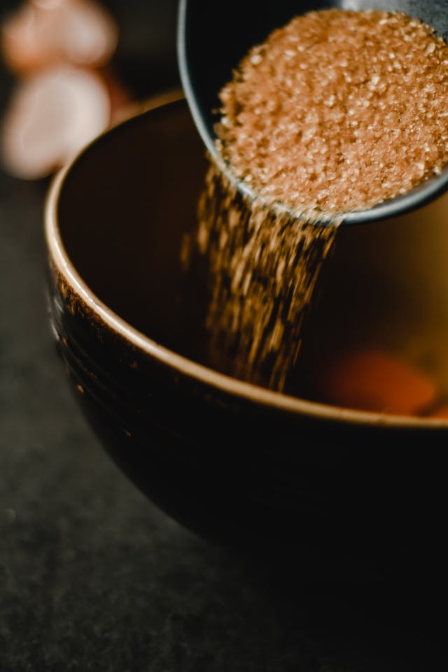 Brown sugar being poured from a measuring cup into a dark ceramic bowl.
