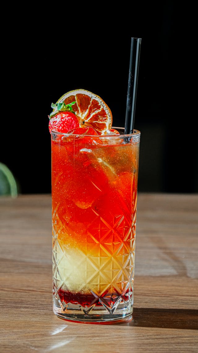Layered red and orange cocktail in a cut-crystal glass, garnished with a dried orange slice and strawberry.