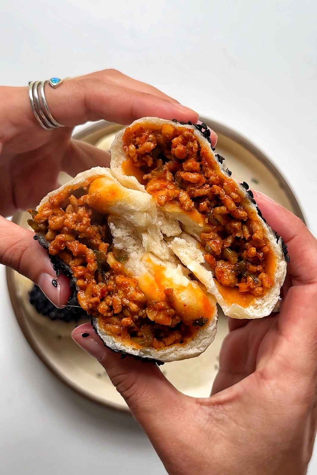 Two hands pulling apart a fluffy white gochujang pork steamed bun showing the spicy red ground pork filling with scallions inside the soft yogurt-and-self-raising-flour dough, sprinkled with black sesame seeds on top.