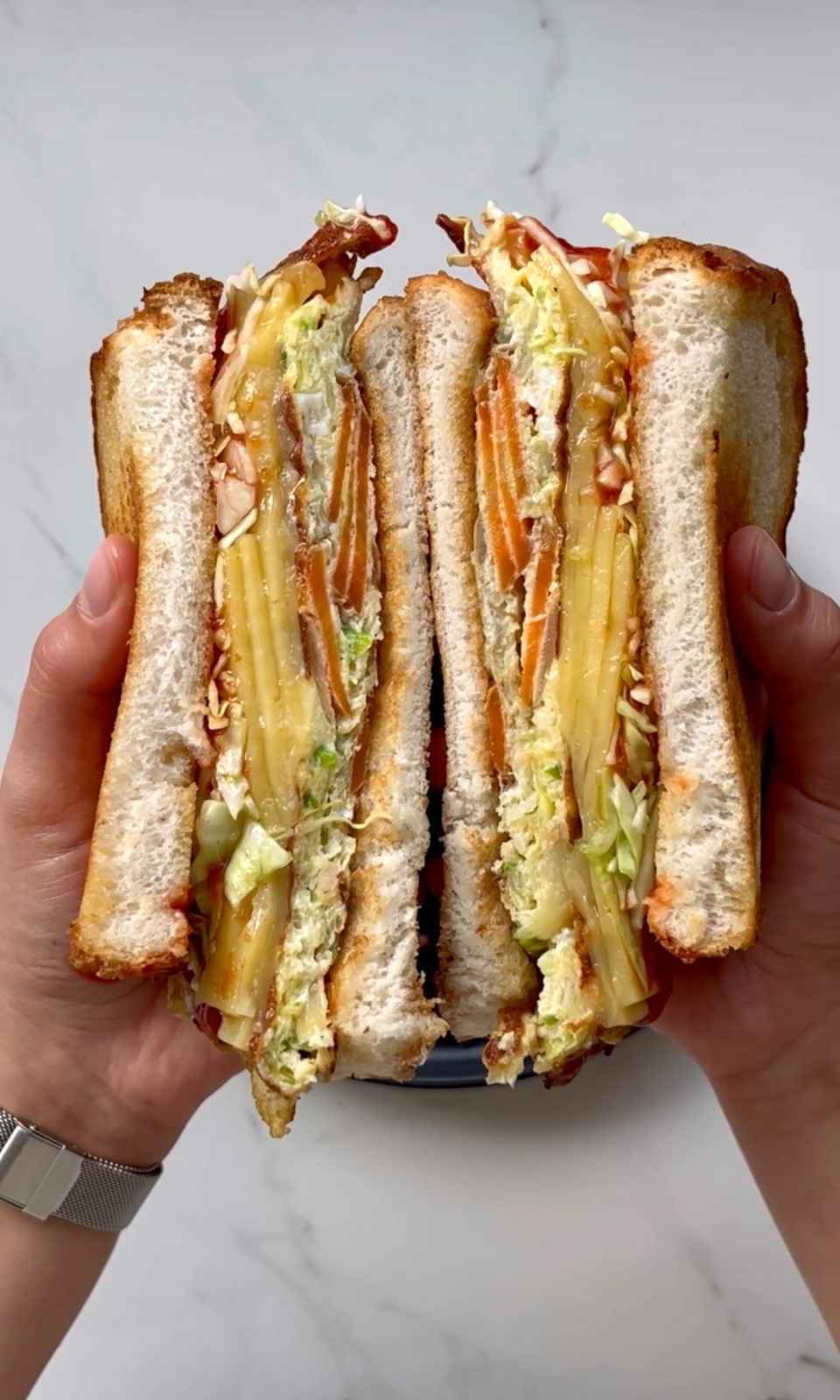 Two hands holding a Korean gilgeori toast sandwich split in half showing a thick veggie omelette filling with shredded cabbage and carrot, melted cheese, ketchup, and a sprinkle of sugar between two slices of golden butter-fried white bread.